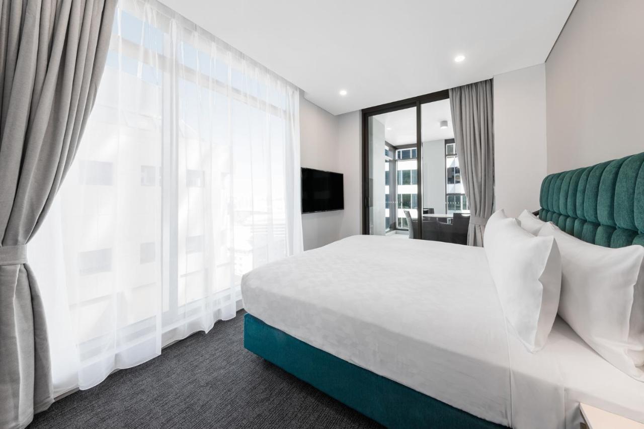 Meriton Suites Sussex Street, Sidney Dış mekan fotoğraf The photo shows a modern bedroom featuring a large bed with a white duvet and pillows. The headboard is turquoise, adding a pop of color to the neutral-toned room. There are sheer white curtains covering large windows that let in natural light, creat