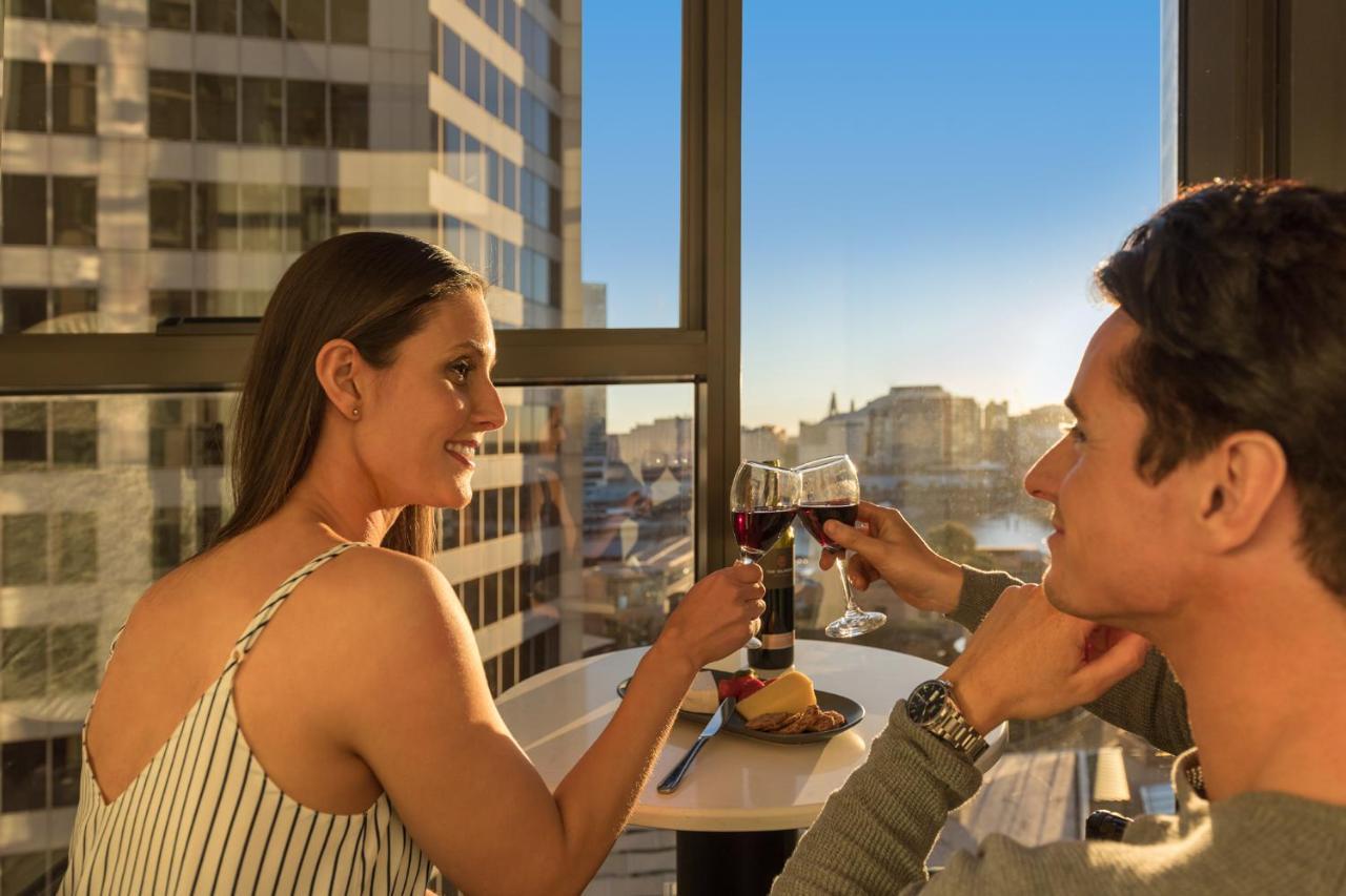 Meriton Suites Sussex Street, Sidney Dış mekan fotoğraf The photo depicts a couple enjoying a drink together in a modern setting. They are sitting at a small table near a large window, which provides a view of a cityscape and a sunset. Both individuals are smiling and engaged in a cheerful toast with glas